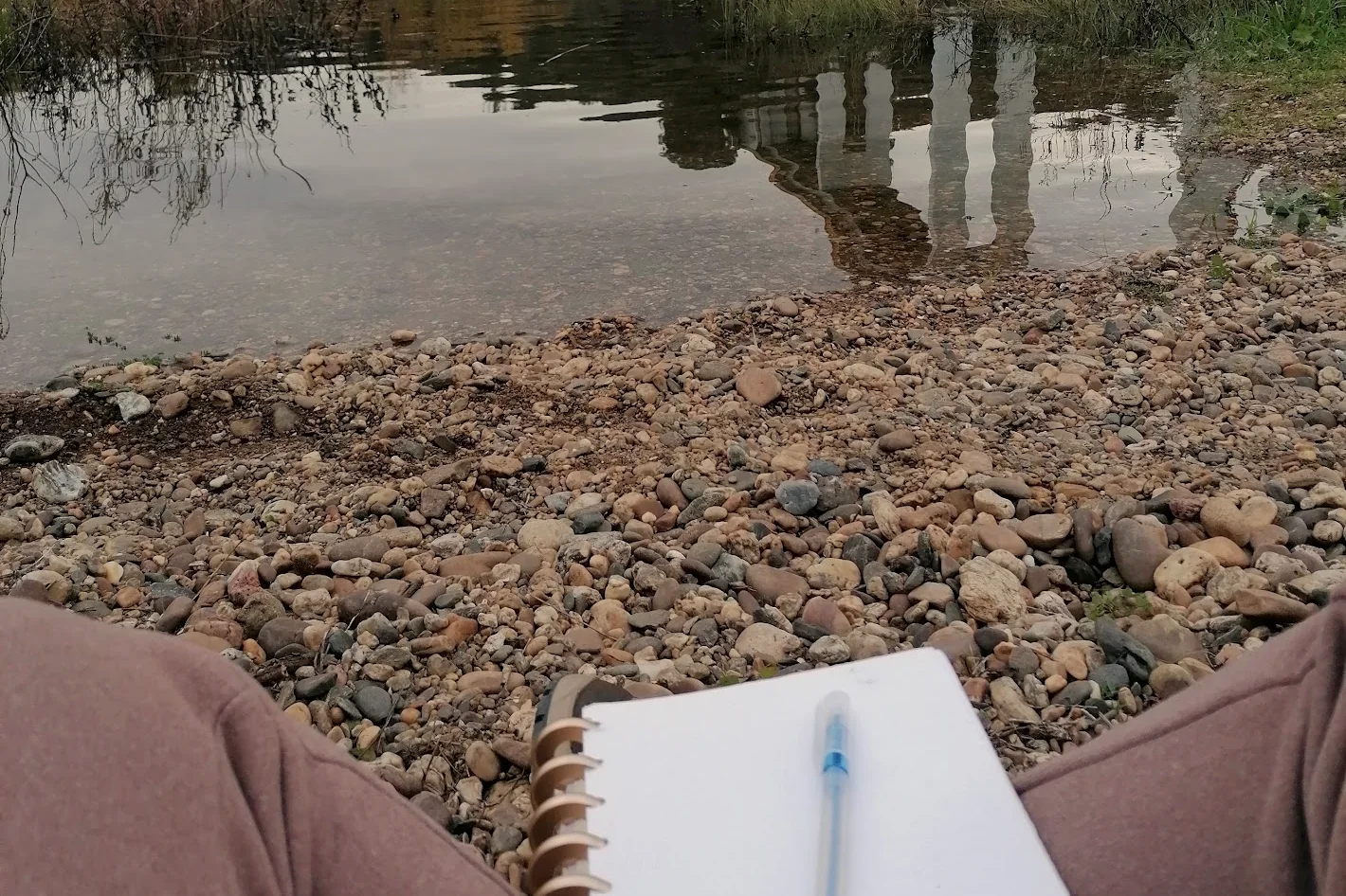 Caderno aberto e caneta pousados sobre os joelhos, à beira de um rio de seixos, num dia encoberto.