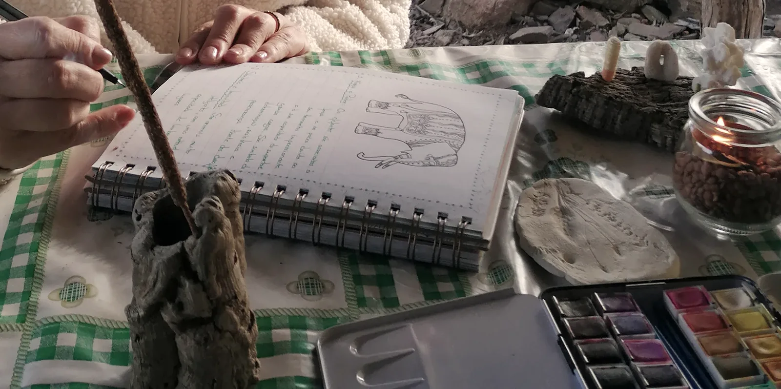 Mãos a desenhar num caderno aberto, com aguarelas, pedras e uma vela sobre uma toalha aos quadrados.