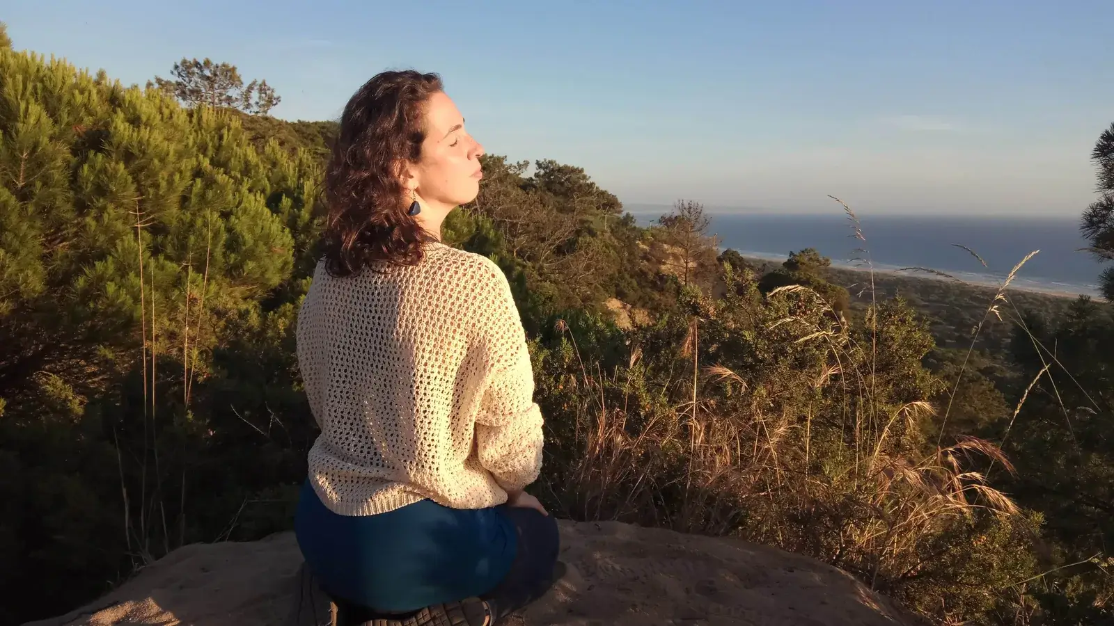 Eugénia Amaro sentada numa rocha, de costas e de perfil, a olhar o pinhal e o mar ao fim da tarde.