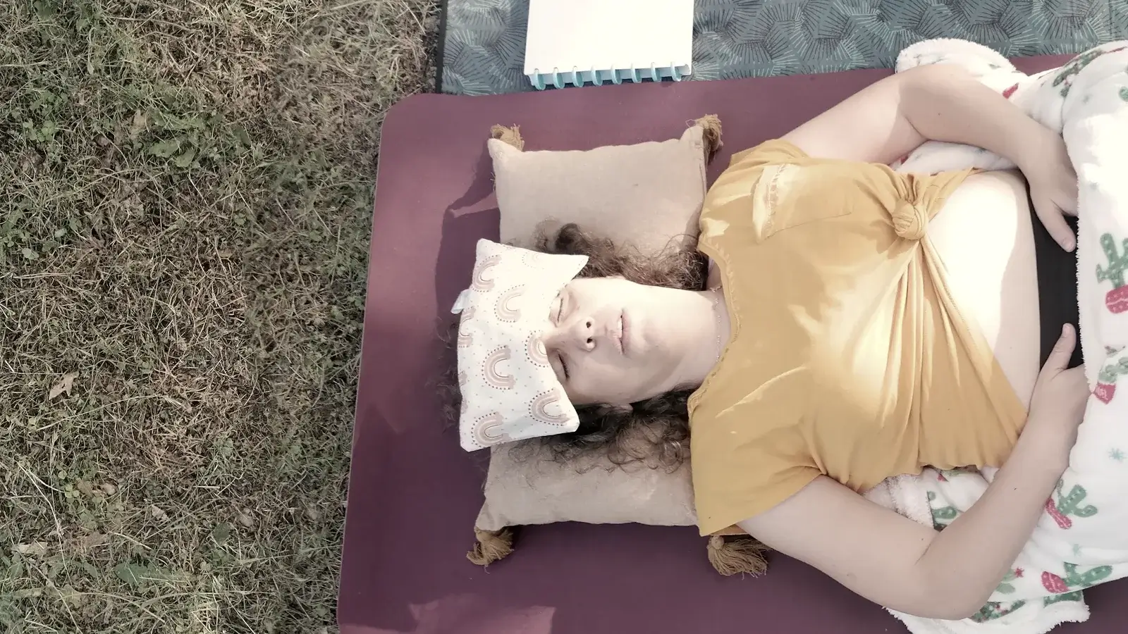 Eugénia Amaro deitada numa postura de Yoga Restaurativo, com almofada de olhos e apoios, sobre um colchão ao ar livre.